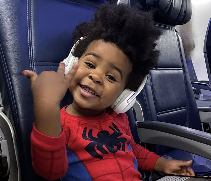 A young boy in a Spider-Man shirt, smiling with headphones on, seated in an airplane.