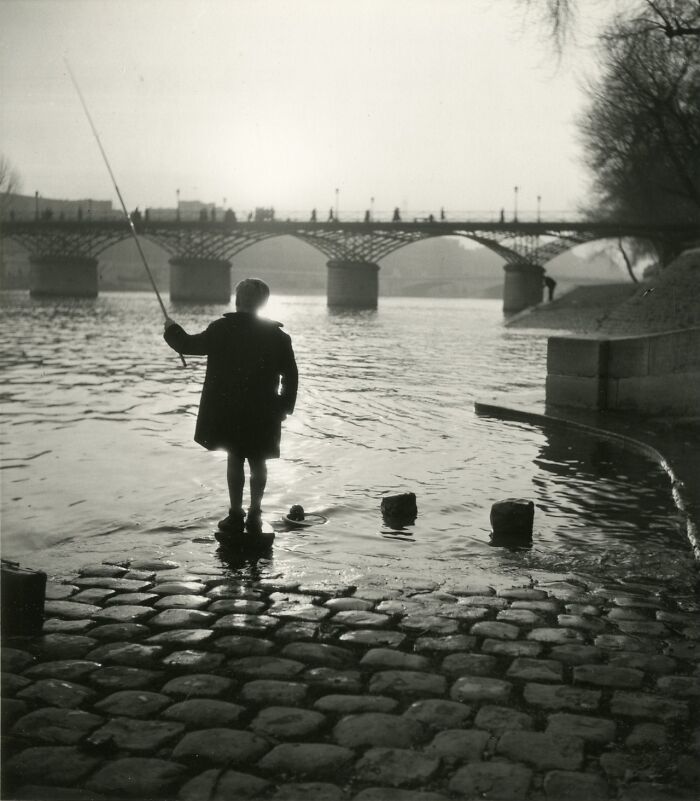 Vintage black and white photo of a child fishing near a river with a historic arched bridge in the background, historical photo.