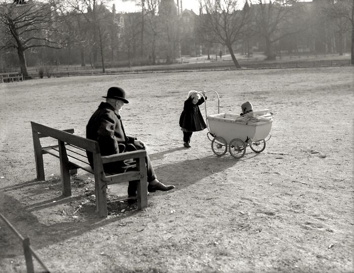 Vintage black and white photo of a man sitting on a bench while a child pushes a baby carriage in a park.