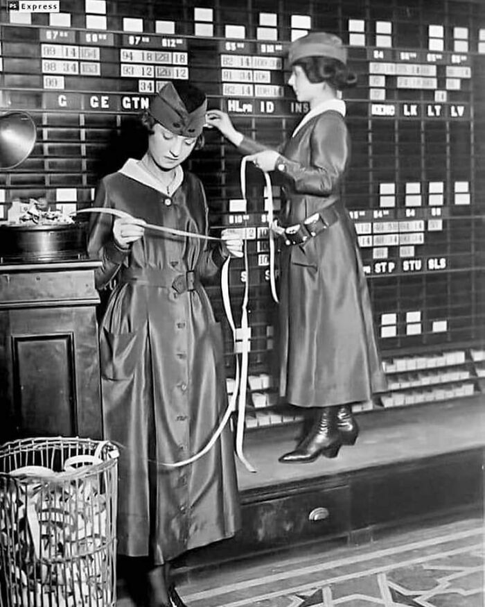 Two women in vintage attire handling long paper tapes in front of a large board, historical and vintage photos.