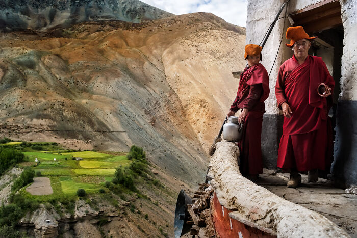 Two monks in traditional robes standing on a balcony overlooking terraced fields in a mountainous region, travel photography award winner.