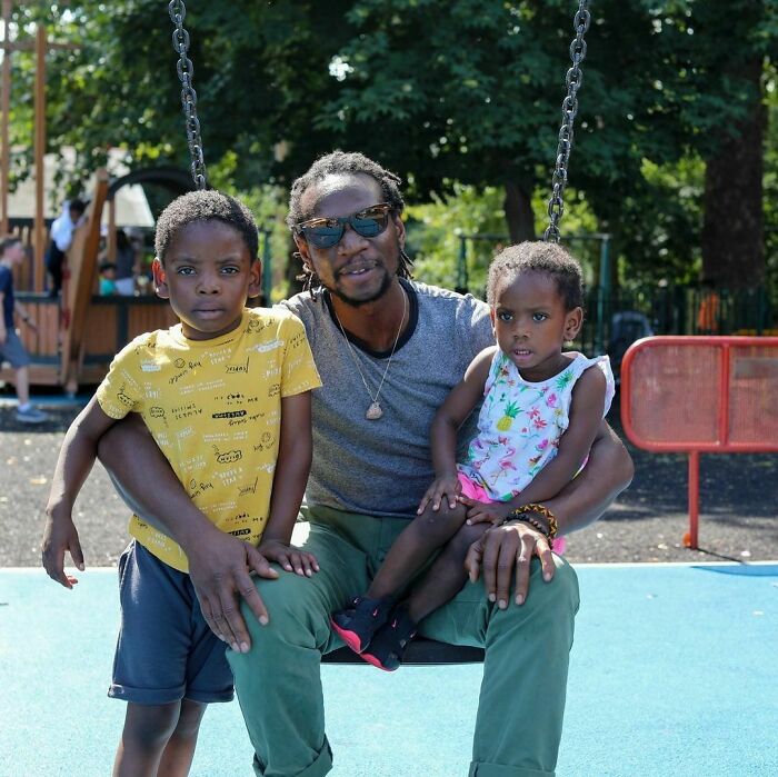 Man wearing sunglasses with two children on a swing at a park, capturing moving stories from Humans of New York.