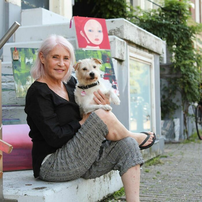 Older woman sitting outdoors holding a small dog, surrounded by colorful artwork, capturing moving stories from Humans of New York.