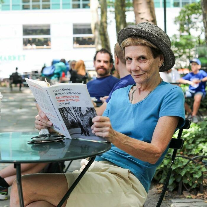 Woman wearing a hat and blue shirt sitting outdoors, reading a book, part of moving stories from Humans of New York.