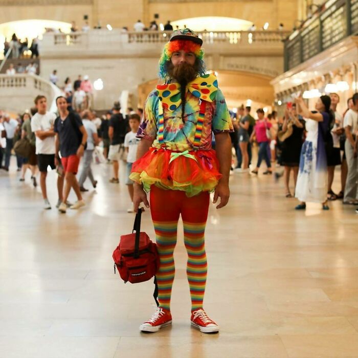 Man in colorful clown outfit with rainbow wig and striped leggings standing in busy public space, humans of New York stories.