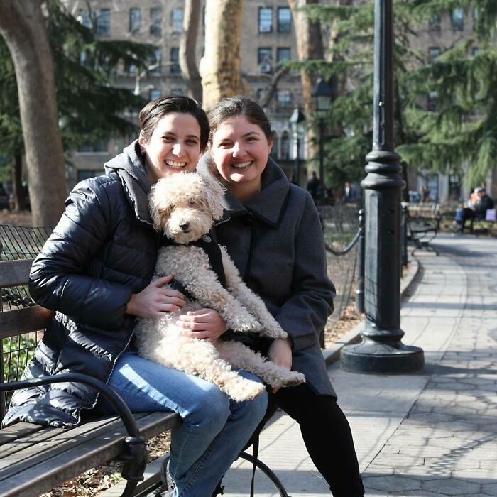 Two women sitting on a park bench in New York, smiling and holding a fluffy dog, capturing moving stories from humans.