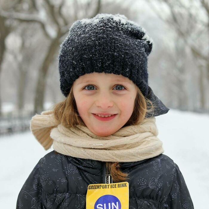 Young girl bundled in winter clothes with snow on her hat, smiling in a snowy park, featured in moving stories Humans of New York.