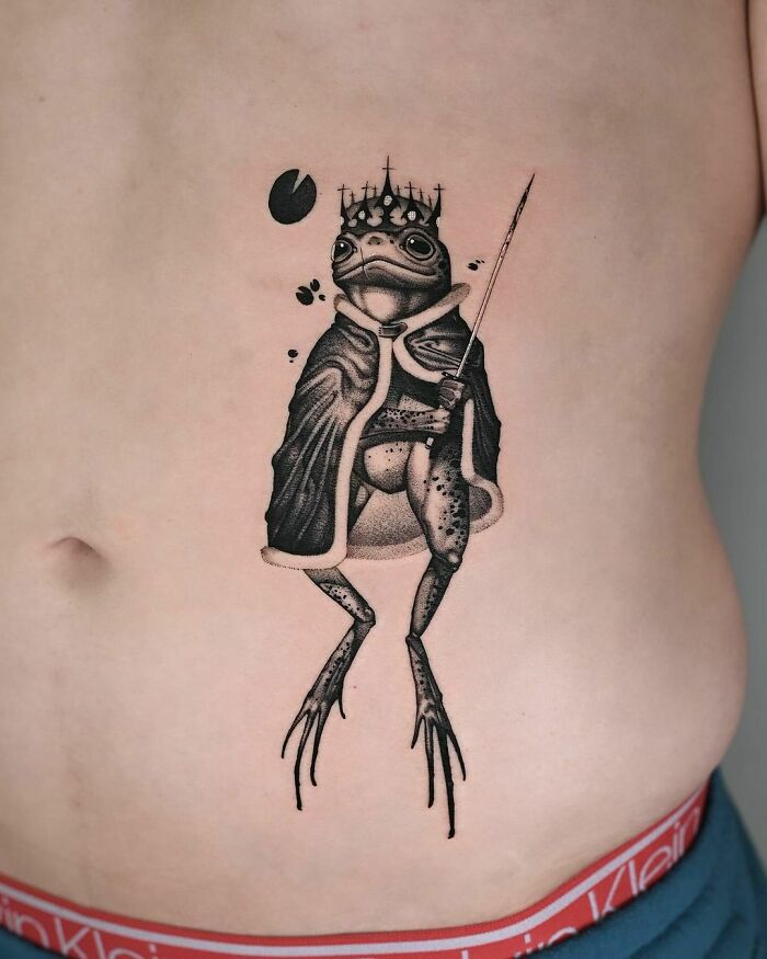 A creative frog tattoo with a royal theme, featuring a crowned frog in a cloak holding a scepter.