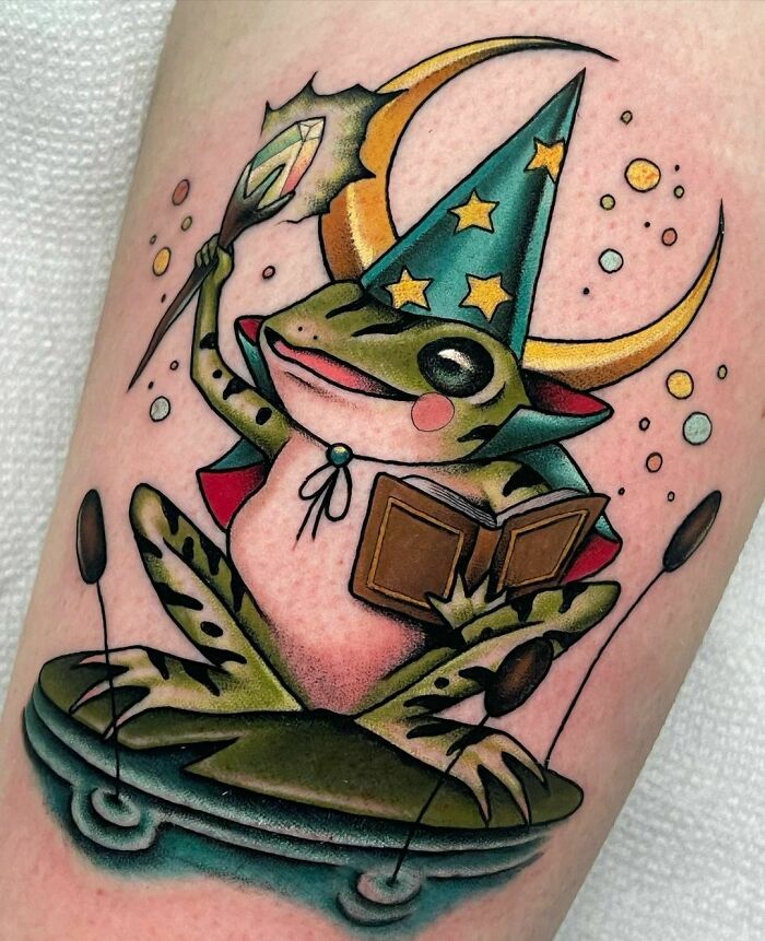 Colorful wizard frog tattoo reading a book, wearing a starry hat, crescent moon in the background.