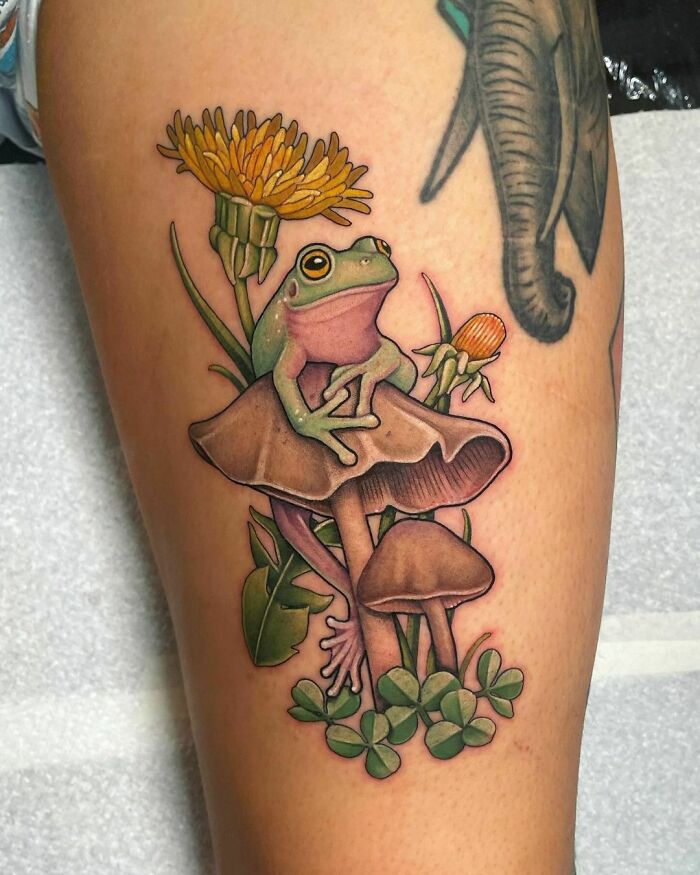 Creative frog tattoo featuring a green frog sitting on mushrooms with dandelions and clovers.