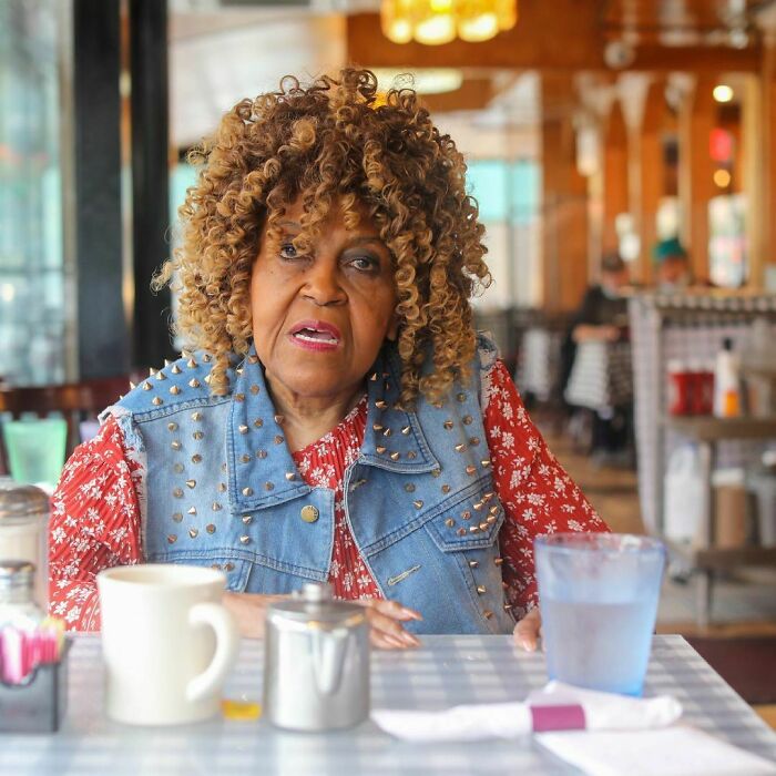 Elderly woman with curly hair sharing one of the most moving stories from Humans of New York in a cafe setting.