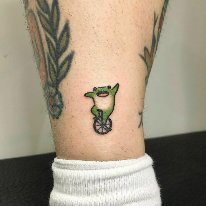 Frog tattoo on a leg, featuring a cartoon frog on a unicycle, captured in a simple and cute style.
