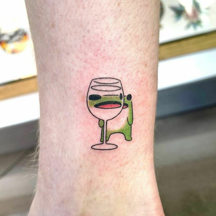 Cartoon frog tattoo with a wine glass on ankle.