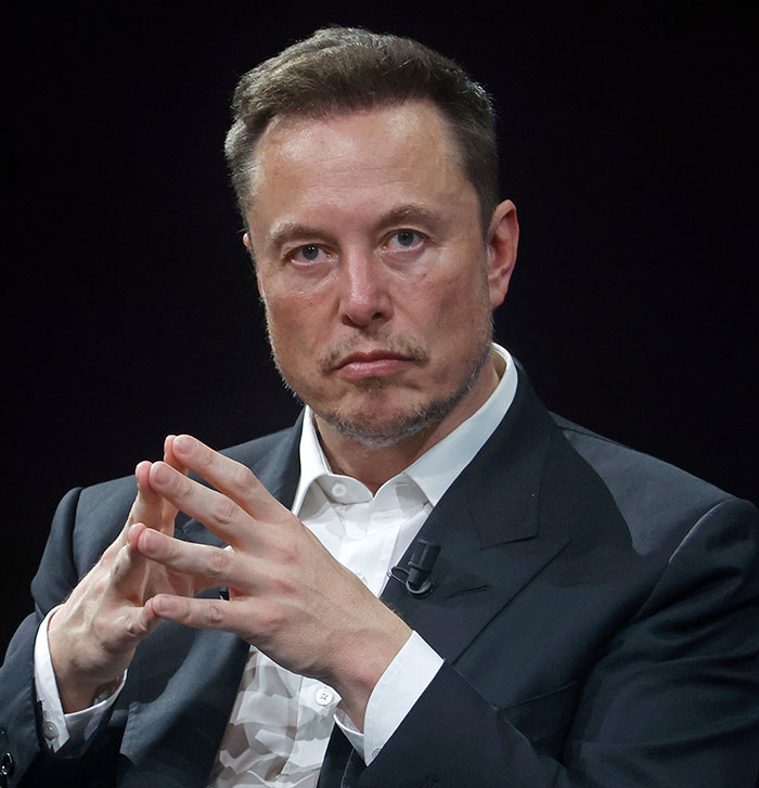 Elon Musk in a dark suit, concentrating with hands clasped during a public event.