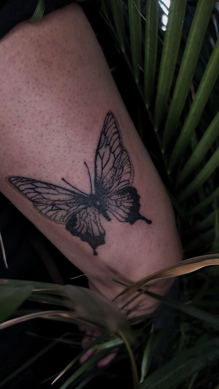 Handpoke tattoo of a butterfly on skin amidst green leaves, showcasing unique artistry.