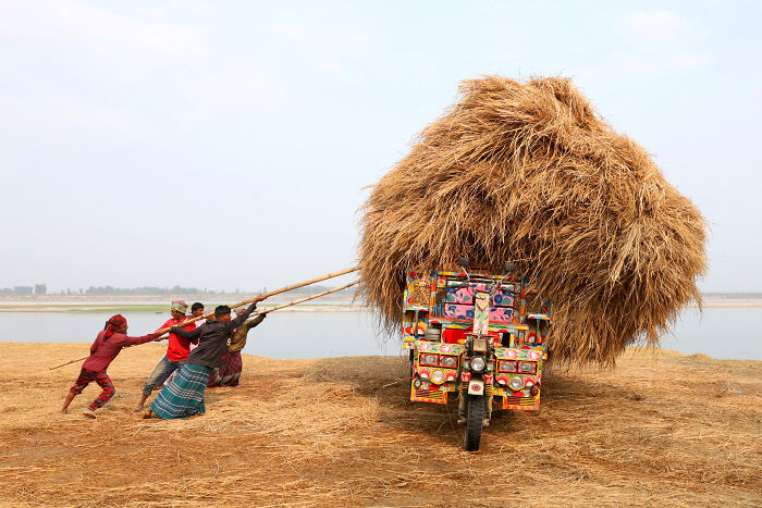 Group of people loading a colorful vehicle with a massive stack of hay near a river, showcasing breathtaking travel photography awards.