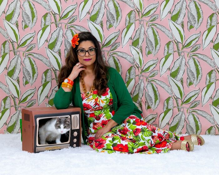 Woman in floral dress and green cardigan poses with her cat inside a vintage TV in quirky pet and owner portraits.