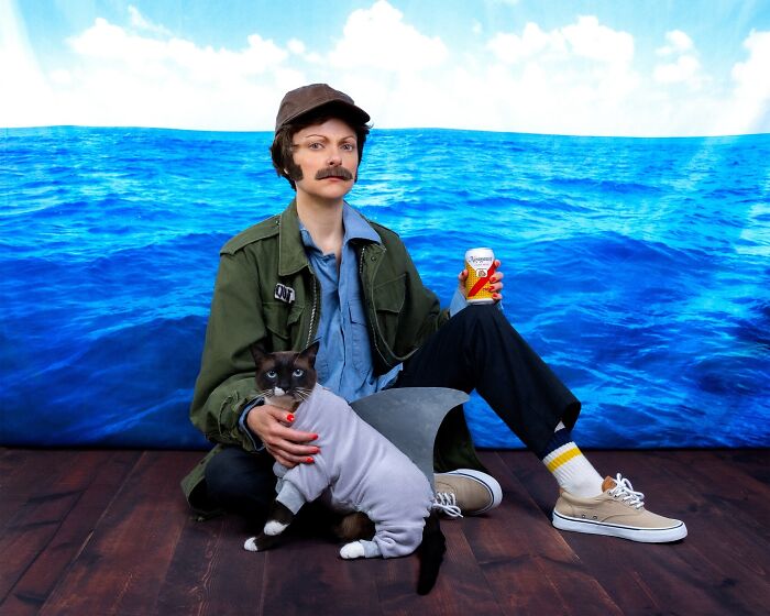 Person with mustache and cap holding a can, sitting beside a cat dressed as a shark in a quirky pet portrait.