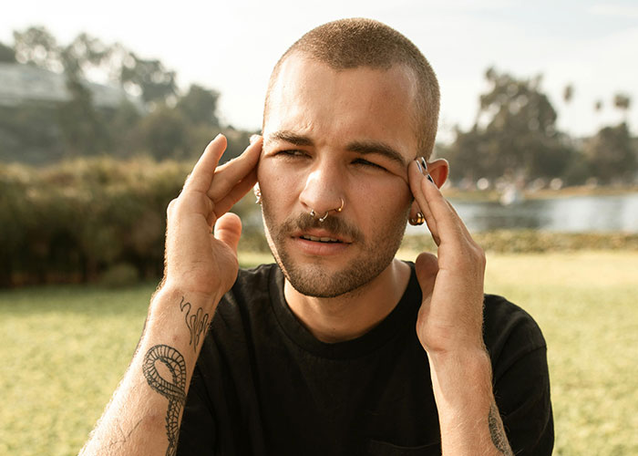 Young adult man with tattoos outdoors, appearing stressed and rubbing his temples, illustrating common adult problems.