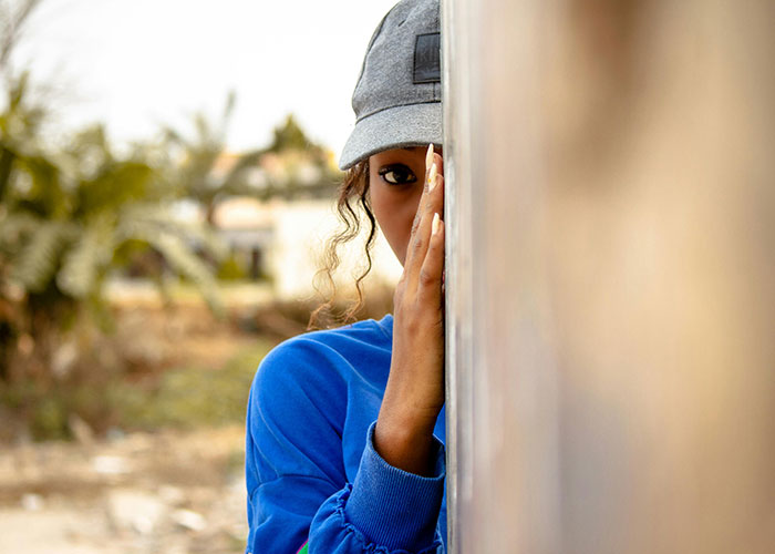 Young adult woman wearing a cap peeking from behind a wall, illustrating common adult problems people face in life.