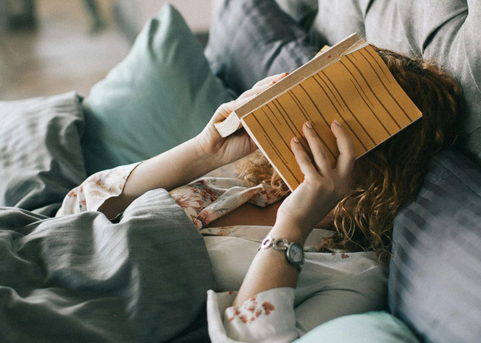 Woman lying in bed with a book covering her face, illustrating adult problems people wish they knew earlier in life.