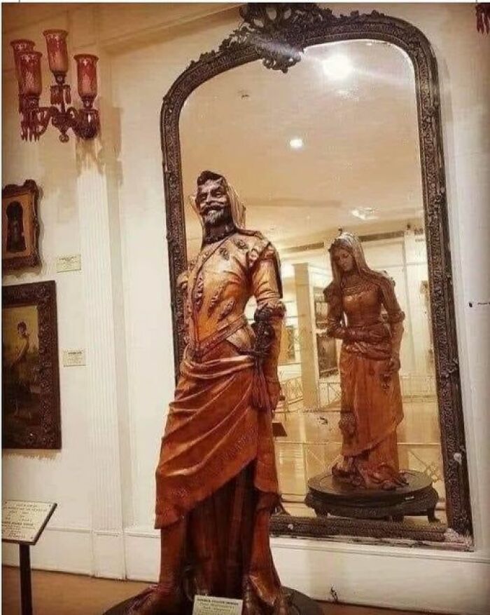 Ancient marvel: intricate wooden statue reflecting in an ornate mirror, detailed craftsmanship on display in a gallery.