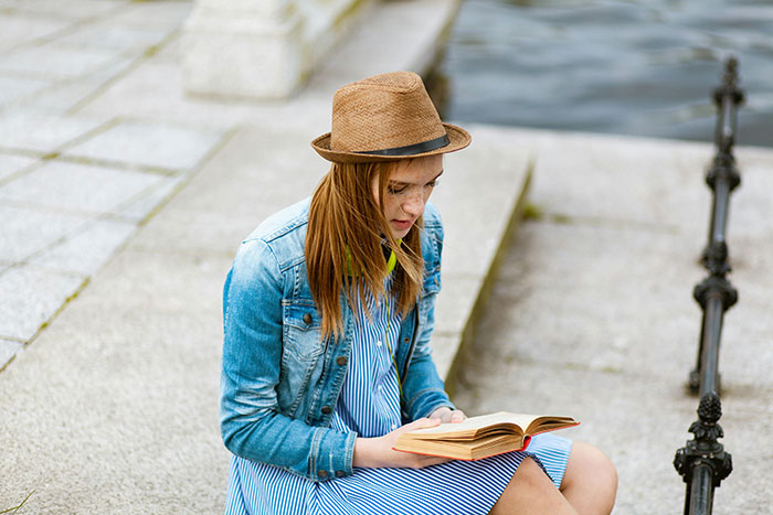 Young woman in a hat and denim jacket reading a book by waterfront, reflecting on stories about the world's worst parents.