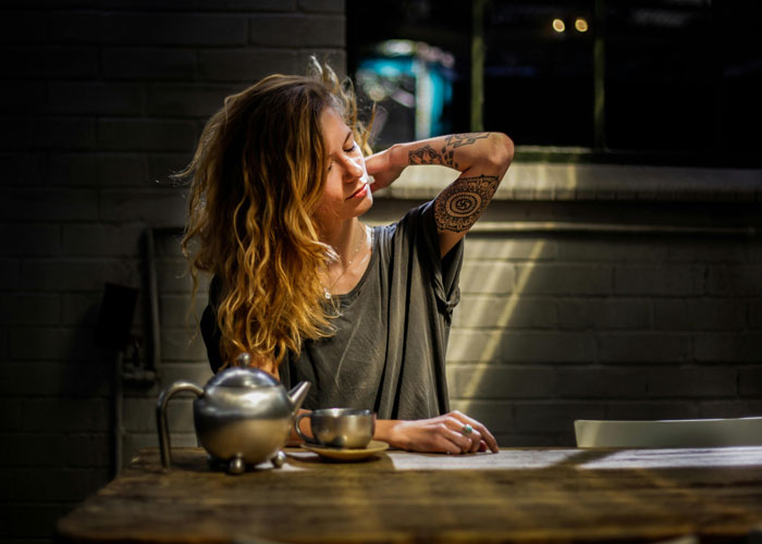 Woman in a dimly lit room, revealing her arm tattoo while sitting at a table with a teapot and cup nearby.