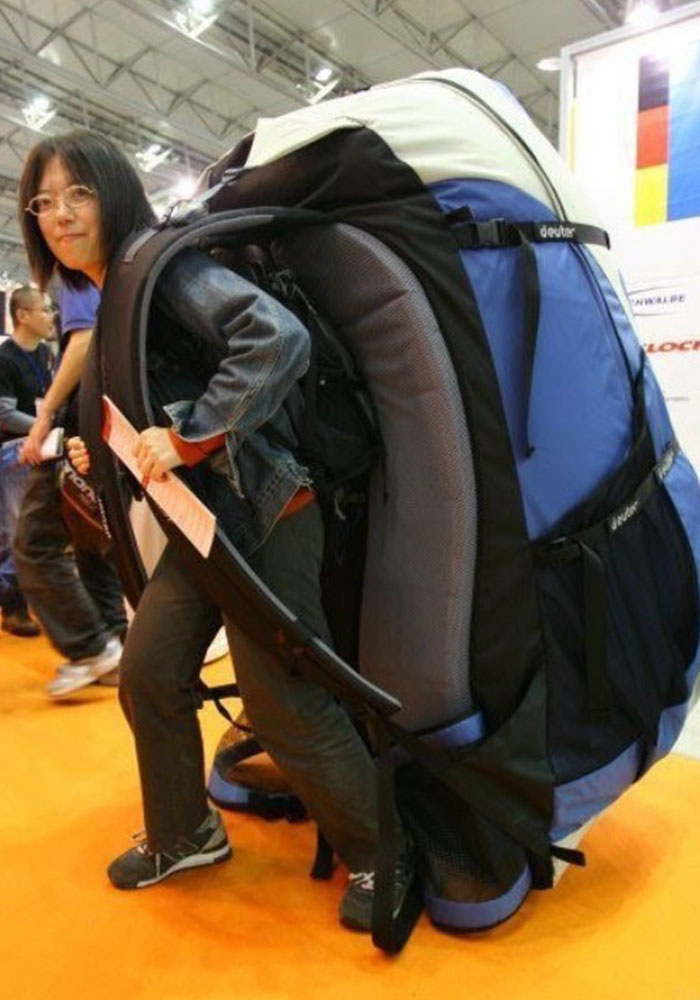  A person wearing an absurdly oversized backpack, which is so large that it dwarfs them, making it appear comically impractical for normal use. The person is leaning forward slightly as if struggling under the weight of the giant pack.