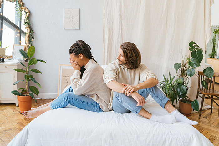 Couple sitting back-to-back on bed, displaying signs of falling out of love in a bright, plant-filled room.