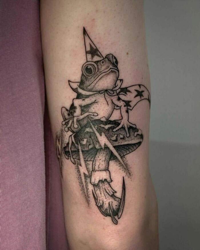 Creative frog tattoo depicting a frog in a wizard hat sitting on a mushroom, designed with stars and lightning motifs.