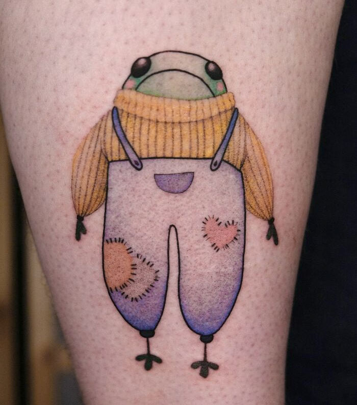 Cute frog tattoo wearing a yellow sweater and colorful overalls, featuring heart patches on the pants.