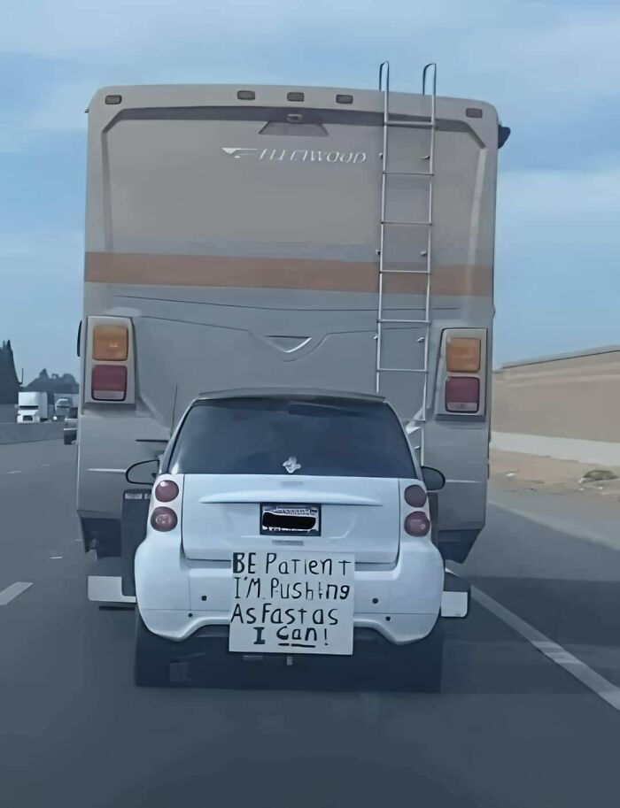 Car meme with a funny sign on a small car behind an RV, saying "Be patient, I'm pushing as fast as I can."