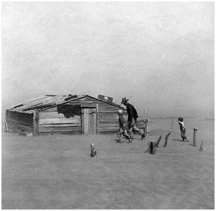 Vintage historical photo of a man and children walking near a weathered shack in a barren, dusty landscape.