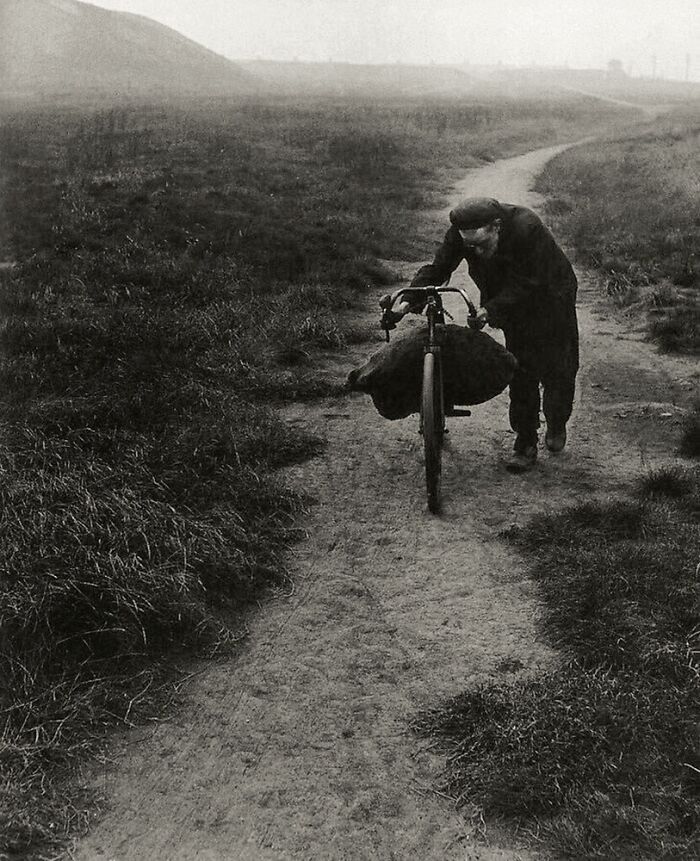 Old black and white vintage photo of a man pushing a bicycle along a dirt path in a rural historical setting