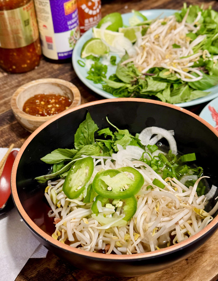 A fresh lunch idea featuring a bowl of Vietnamese pho, garnished with bean sprouts, jalapeño slices, basil, and onions. In the background, a plate with additional herbs, lime wedges, and condiments is visible alongside sauces and chili paste. A fresh lunch idea featuring a bowl of Vietnamese pho, garnished with bean sprouts, jalapeño slices, basil, and onions. In the background, a plate with additional herbs, lime wedges, and condiments is visible alongside sauces and chili paste.