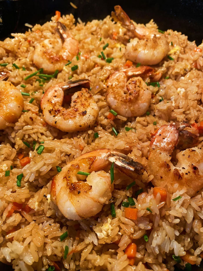 A savory lunch idea featuring fried rice with grilled shrimp, garnished with chopped chives and small pieces of carrots, served in a skillet. A savory lunch idea featuring fried rice with grilled shrimp, garnished with chopped chives and small pieces of carrots, served in a skillet.