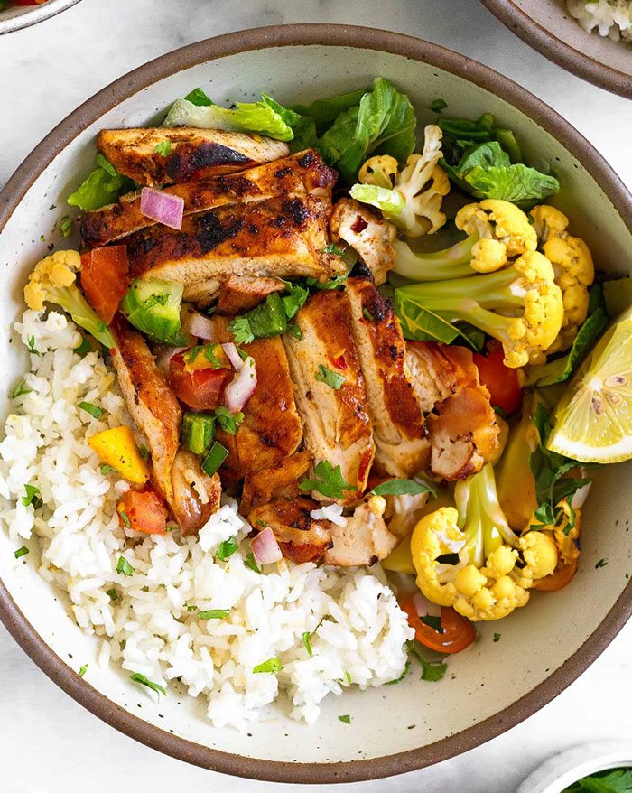 A healthy lunch idea featuring grilled chicken slices served over a bed of white rice, accompanied by roasted cauliflower, fresh lettuce, diced vegetables, and a lemon wedge, all arranged in a bowl. A healthy lunch idea featuring grilled chicken slices served over a bed of white rice, accompanied by roasted cauliflower, fresh lettuce, diced vegetables, and a lemon wedge, all arranged in a bowl.