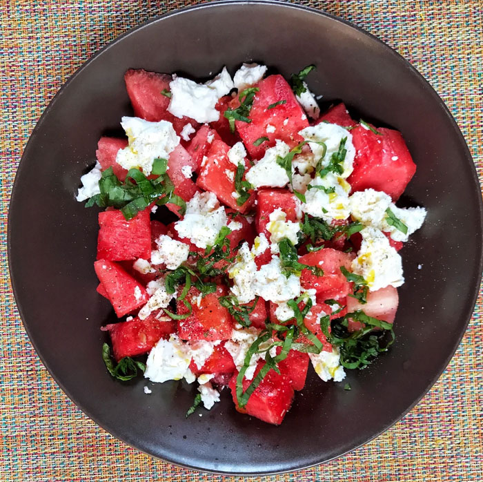A refreshing lunch idea featuring a watermelon salad with crumbled feta cheese, fresh mint, and a drizzle of olive oil, served in a dark bowl on a colorful woven mat. A refreshing lunch idea featuring a watermelon salad with crumbled feta cheese, fresh mint, and a drizzle of olive oil, served in a dark bowl on a colorful woven mat.