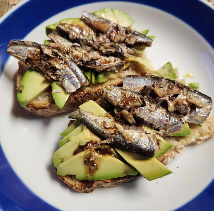 A simple lunch idea featuring toasted bread topped with slices of avocado and sardines, drizzled with a light sauce, served on a white and blue plate. A simple lunch idea featuring toasted bread topped with slices of avocado and sardines, drizzled with a light sauce, served on a white and blue plate.