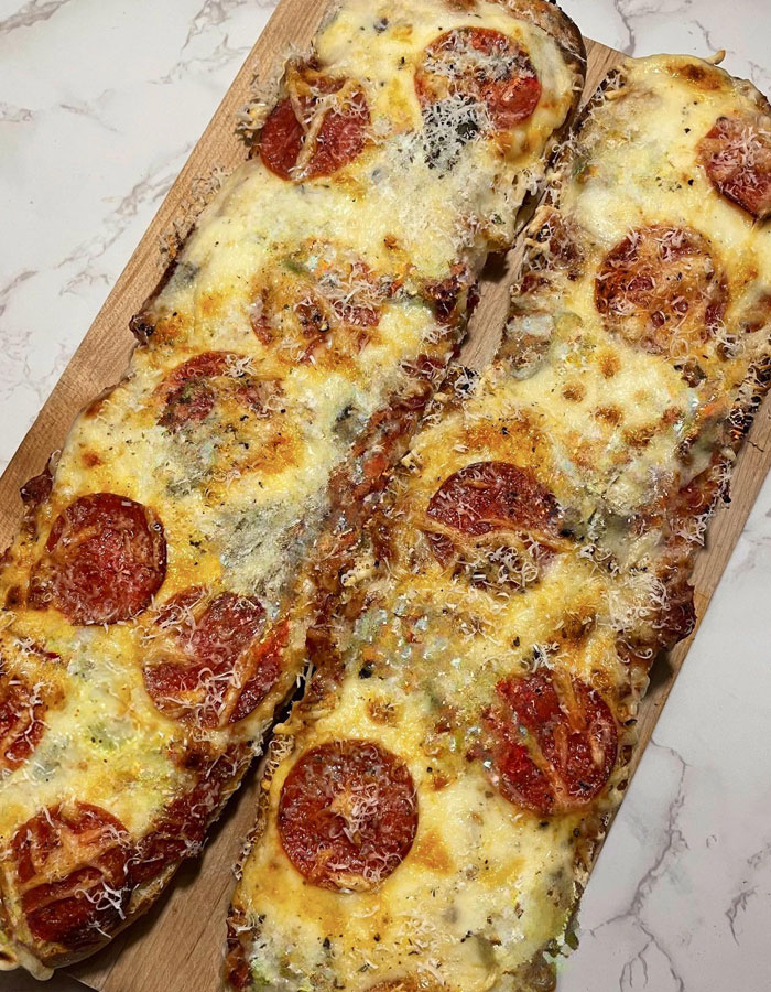 Lunch idea featuring two long flatbread garlic bread pizzas topped with pepperoni, melted cheese, and grated Parmesan on a wooden serving board placed on a marble surface. Lunch idea featuring two long flatbread garlic bread pizzas topped with pepperoni, melted cheese, and grated Parmesan on a wooden serving board placed on a marble surface.