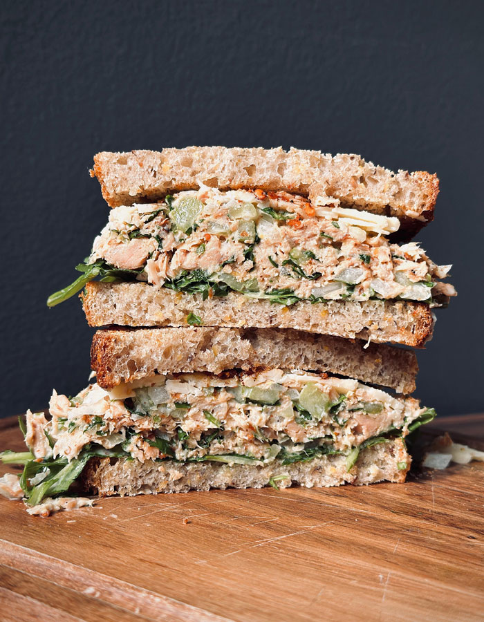 A Lunch idea featuring a hearty sandwich with tuna salad, greens, and vegetables stacked between two slices of whole grain bread on a wooden surface. A Lunch idea featuring a hearty sandwich with tuna salad, greens, and vegetables stacked between two slices of whole grain bread on a wooden surface.