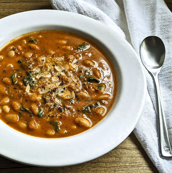 A lunch idea featuring a hearty bowl of bean soup with a rich tomato-based broth, topped with grated cheese and pepper, served with a spoon on the side. A lunch idea featuring a hearty bowl of bean soup with a rich tomato-based broth, topped with grated cheese and pepper, served with a spoon on the side.