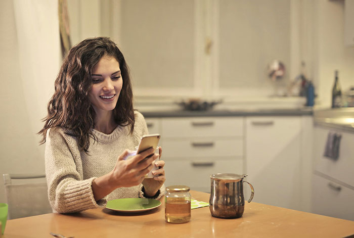 Young woman with natural charm smiling at phone while sitting at kitchen table, showcasing minor attractive features on women.