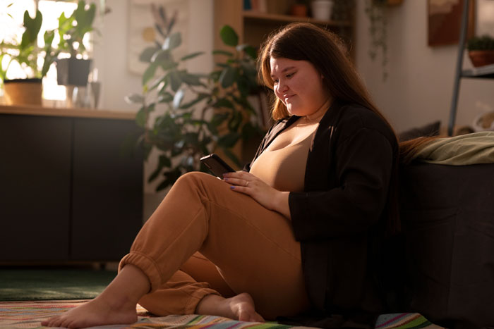 Obese person relaxing at home, using a smartphone with plants in the background.
