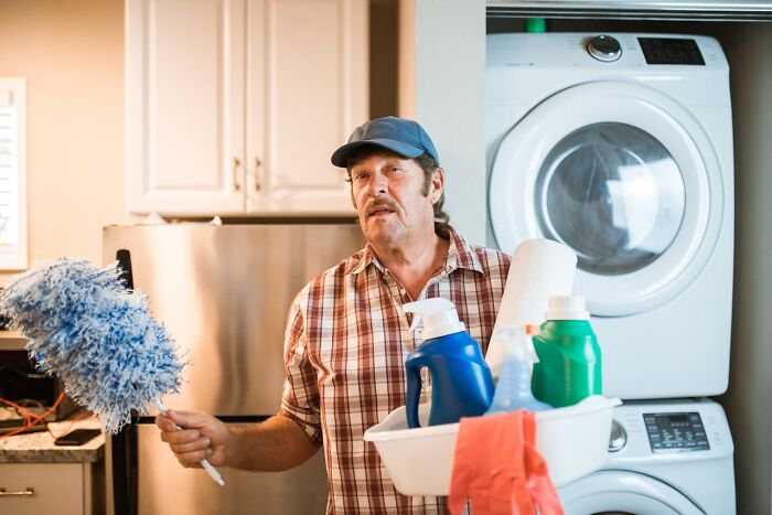 Man holding cleaning supplies and duster in laundry room, illustrating things women want men to understand about chores.