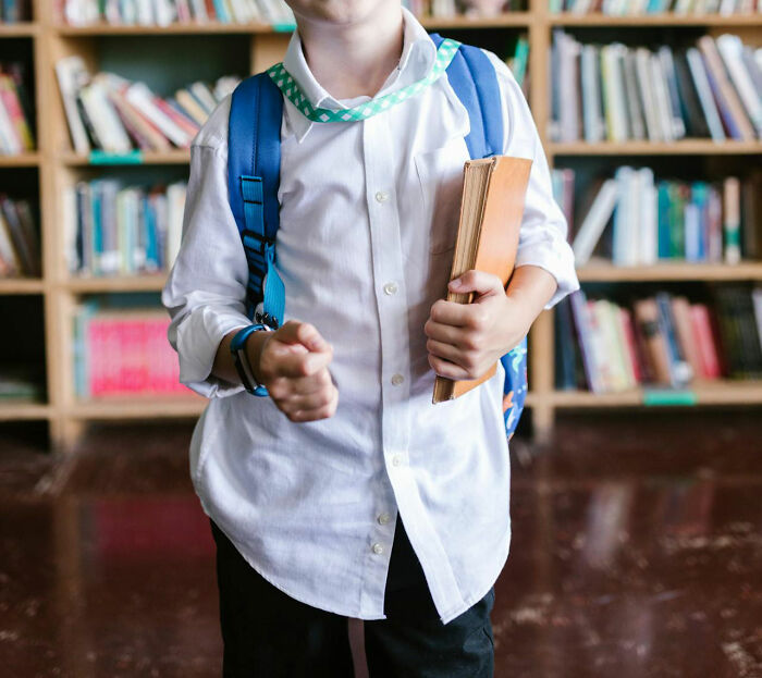 Child in white shirt and backpack holding a book in a library, illustrating white lies backfiring in everyday situations.