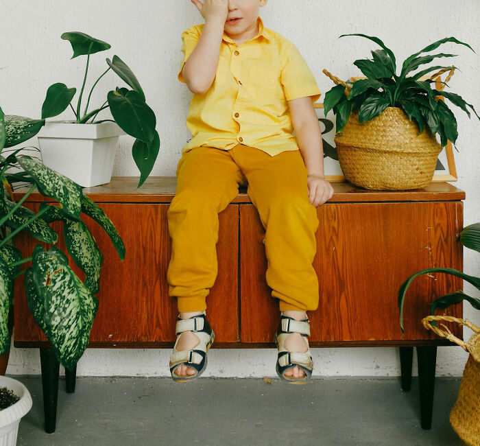 Child in yellow outfit sitting on a wooden cabinet, surrounded by green plants, illustrating white lies backfiring concept.