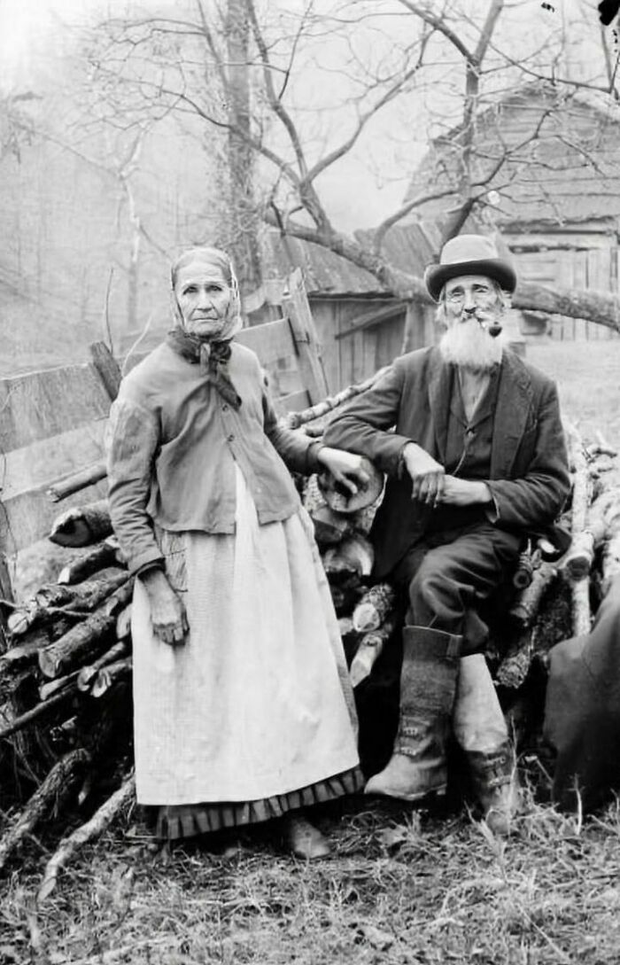 Photograph Of An Elderly Man And Woman Wearing Work Clothes And Seated On A Pile Of Firewood