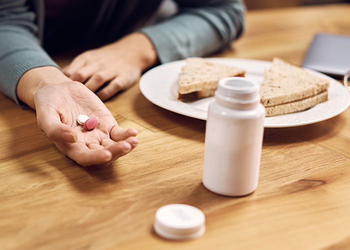 Hand holding pills near a sandwich and a pill bottle on a wooden table representing food thieves at work lessons.